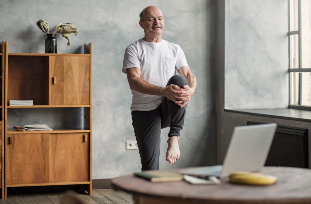 An older adult performing a knee-raise balance exercise and smiling.