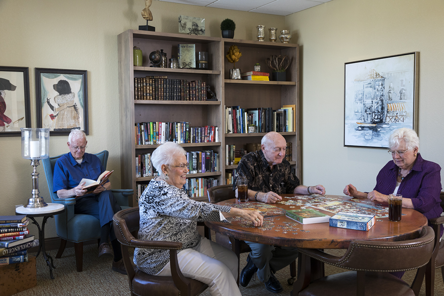 A group of 3 older residents of Rosemark at Mayfair Park builds puzzles as another resident reads a book in the background.