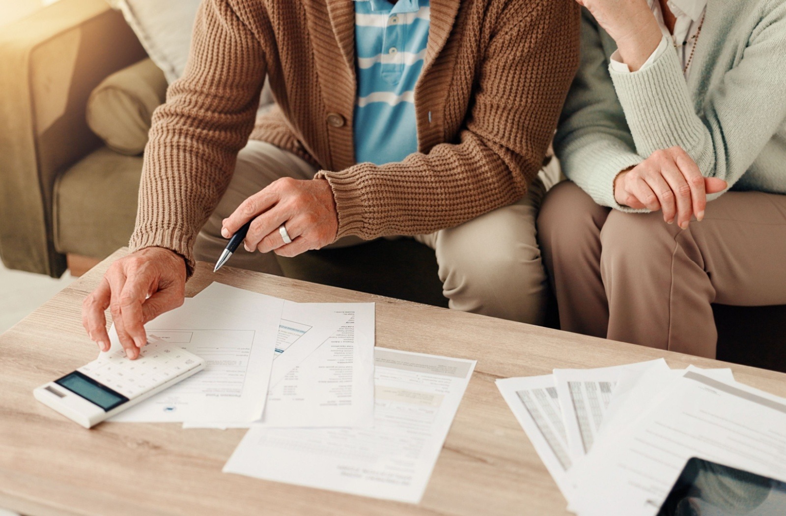 Older man using a calculator while a partner looks on, with financial documents spread across a wooden table.