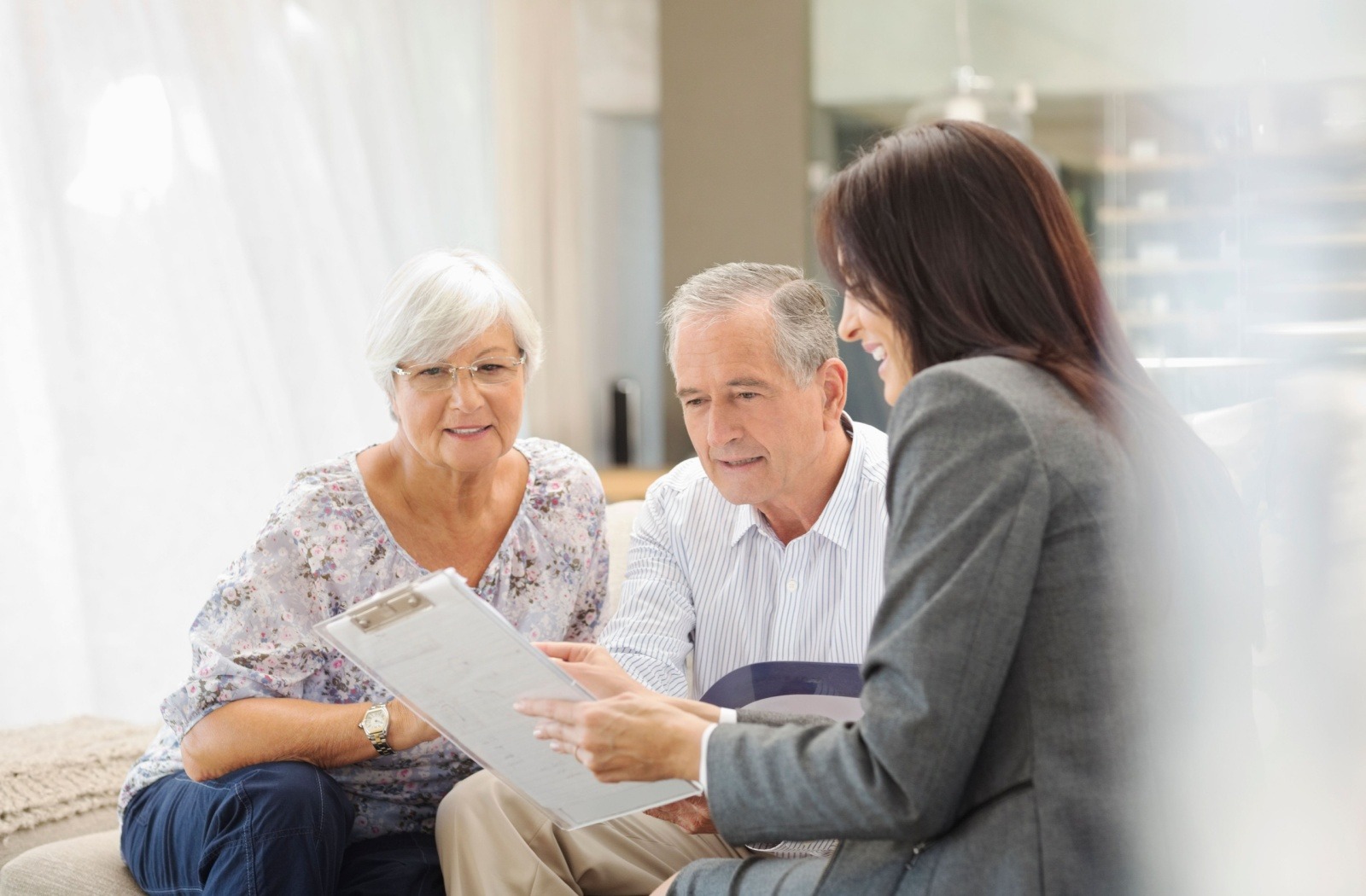 A professional woman in a gray blazer reviews a document with an elderly couple seated on a couch in a bright, modern room.