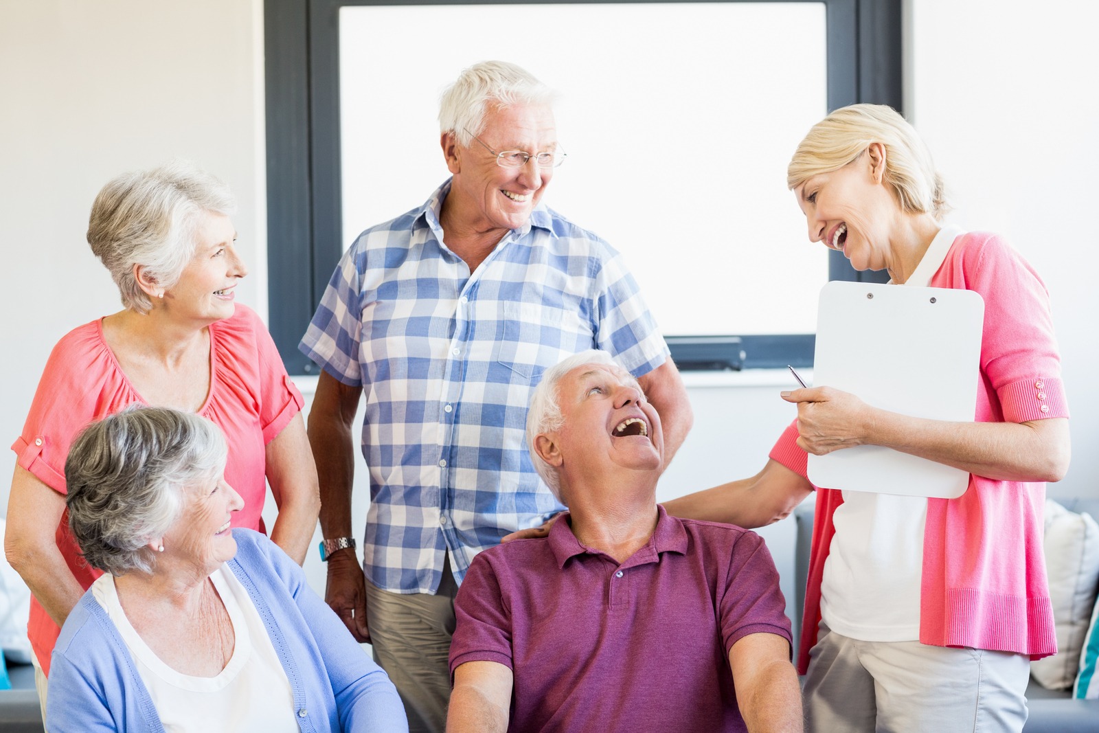 A group of older adults chats with a caregiver while relaxing on a comfortable couch in a senior living apartment, sharing warm smiles.